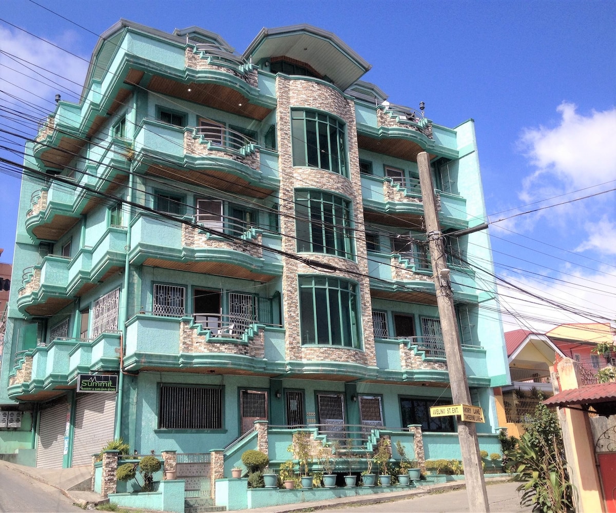 The exterior of the apartelle unit showcases a multi-story turquoise building with stone accents. A landscaped area with potted plants is visible in front of the entrance. Clear skies and bright sunlight illuminate the structure, highlighting its architectural details.