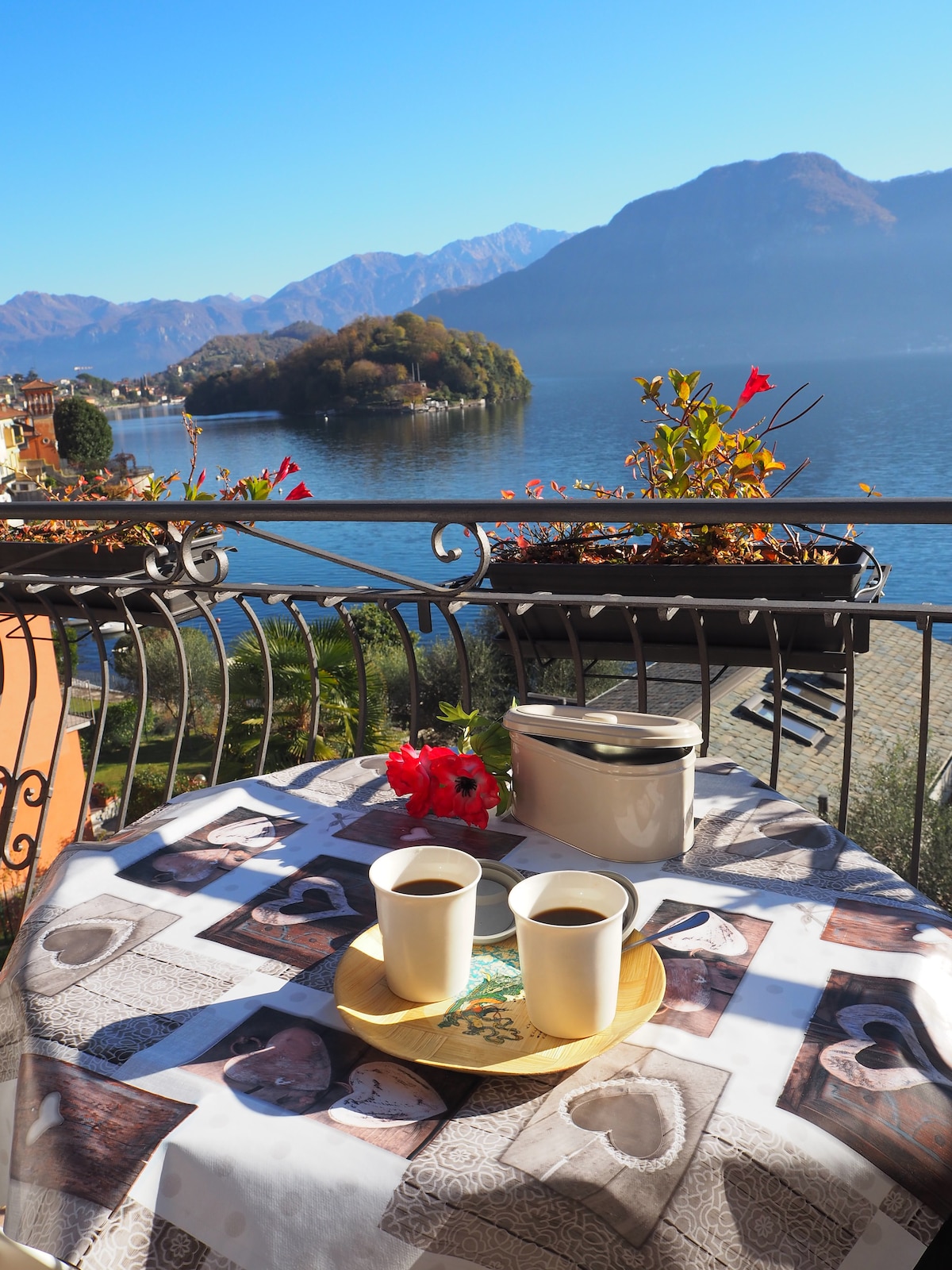 A balcony table is set for two, featuring two cups of coffee on a round wooden tray. The tablecloth displays a design with various patterns. In the background, the serene lake, surrounded by mountains and an island, is visible under a clear blue sky.