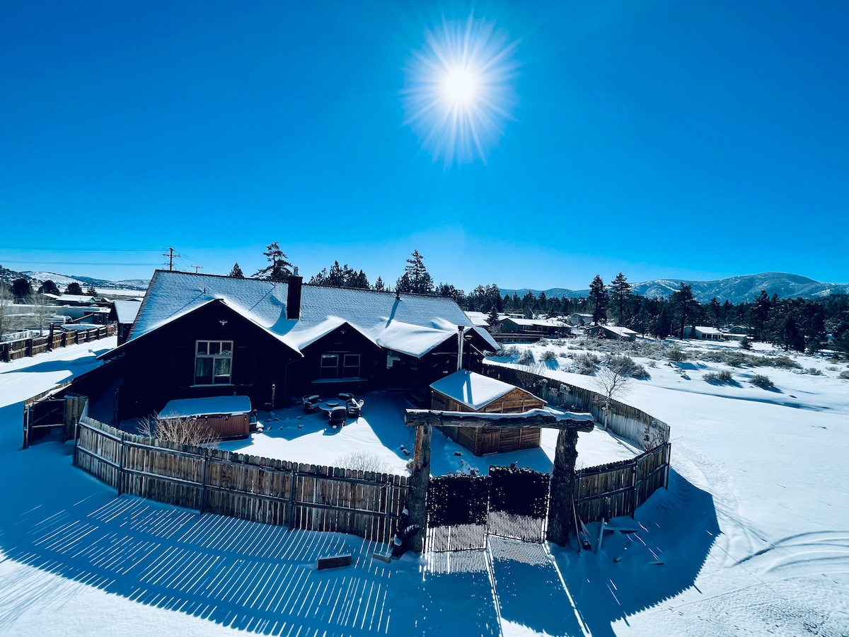 A rustic ranch villa is surrounded by a pristine blanket of snow, with a wooden fence enclosing the property. Clear blue skies and a bright sun highlight the snow-covered landscape, offering a serene winter setting. Tall pine trees are visible in the background.