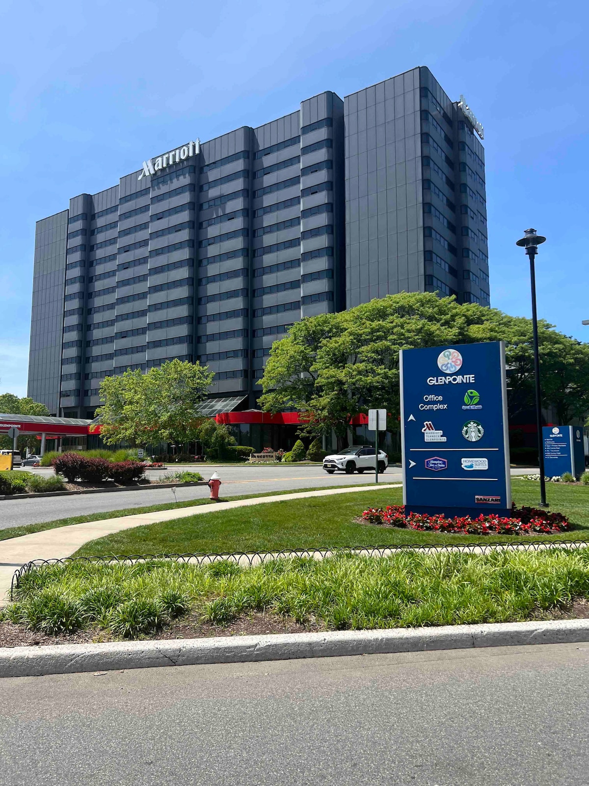 A multi-story hotel building is prominently showcased in the image, featuring a sleek glass façade. Lush greenery surrounds the landscape, with flowering plants and well-maintained grass in the foreground. Informational signage is visible, indicating nearby amenities and services.