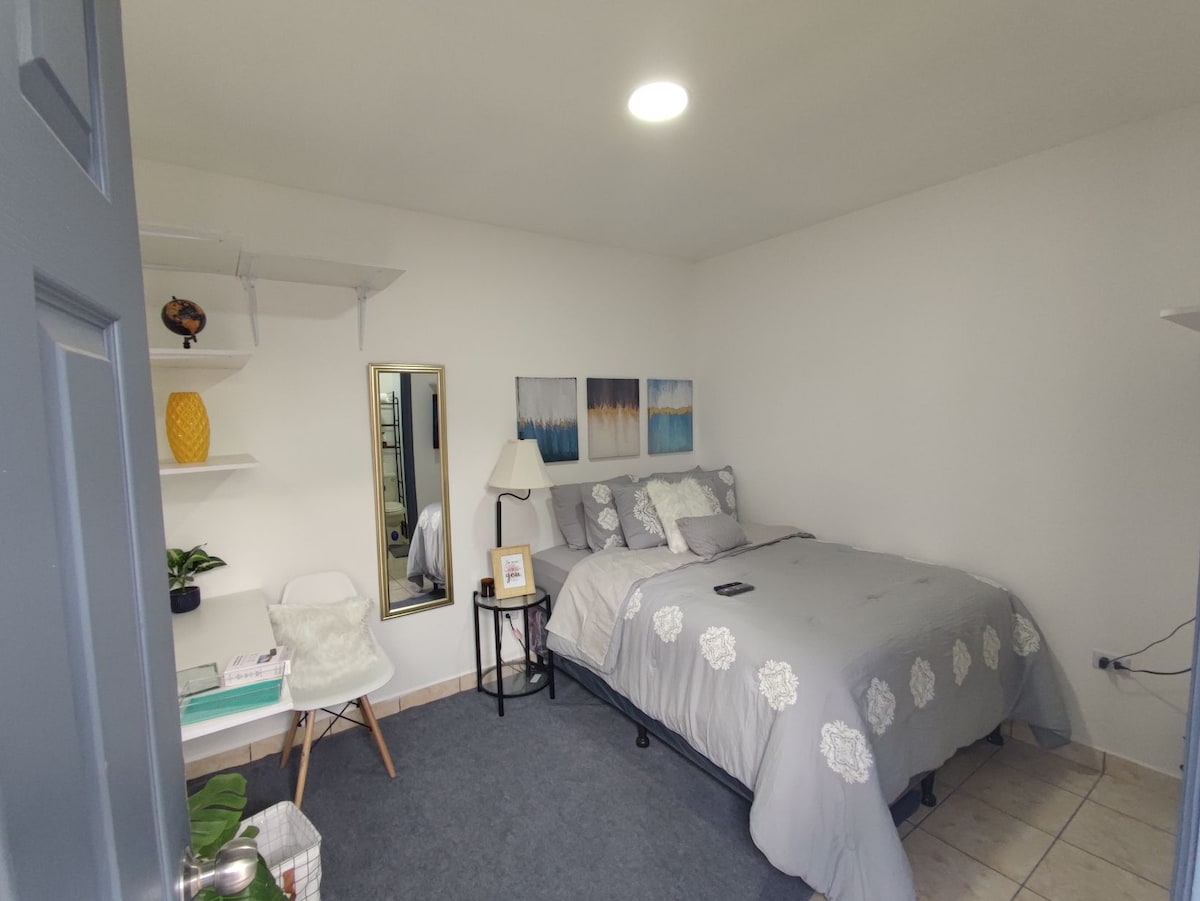 A serene bedroom features a spacious bed adorned with a grey comforter and decorative pillows. A minimalist desk with a chair is positioned beside a mirror. Soft lighting enriches the room, accompanied by subtle wall art and practical shelving displaying decorative items.