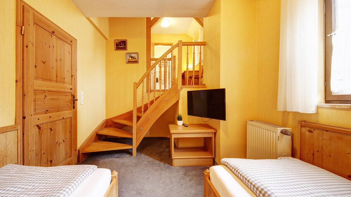 A well-lit room features two beds with patterned bedding positioned next to a wooden nightstand. A television is mounted on the wall, and a modern wooden staircase leads to an upper level. Light-colored curtains allow natural light to illuminate the space.