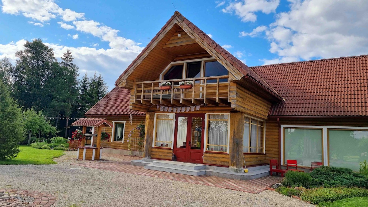 A wooden exterior reflects traditional architecture, featuring a sloped roof and large, decorative windows. A welcoming entrance is highlighted by red doors, flanked by flower boxes. Lush greenery surrounds the property, enhancing the sense of space and tranquility.
