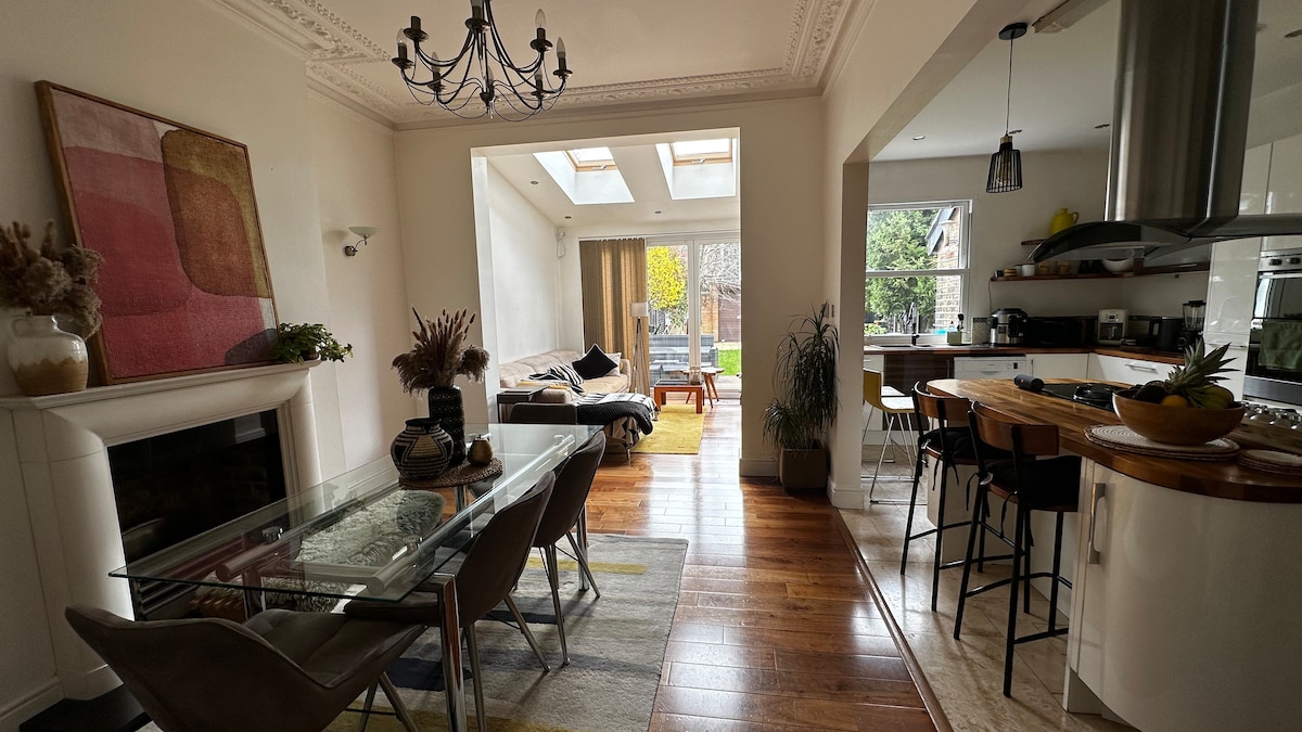 An open-concept living area features a glass dining table surrounded by chairs and a cozy seating arrangement with a sofa. Natural light floods the space through skylights, highlighting the wooden flooring. A kitchen counter with bar stools is visible, creating a seamless flow between dining and kitchen areas.