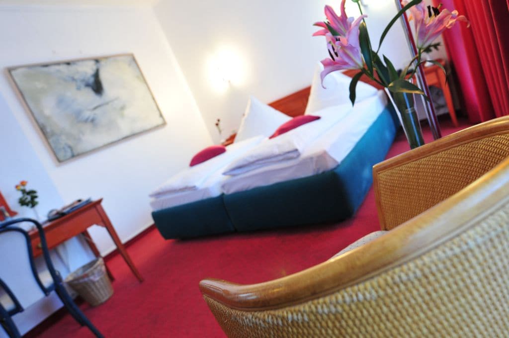 A bedroom features a spacious bed with white linens and decorative cushions in red. A small wooden desk and chair are present, while a potted flower adds a touch of color. The floor is carpeted in red, and a piece of art adorns the wall.