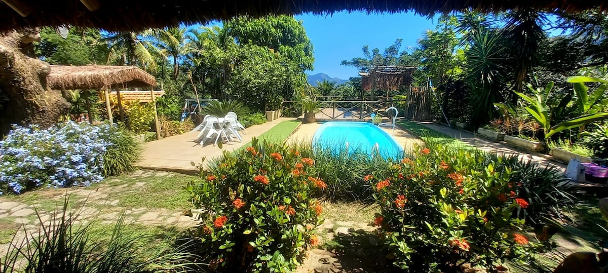 A serene outdoor area showcases a sparkling blue pool surrounded by lush greenery and vibrant flowers. Relaxing seating options are visible, including white lounge chairs. The backdrop features distant mountains, enhancing the tranquil setting amidst various trees and plants.