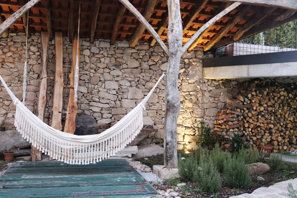 A serene outdoor area features a white hammock suspended between wooden beams under a rustic stone structure. Soft lighting highlights the surrounding greenery and stacked firewood, creating a peaceful atmosphere.