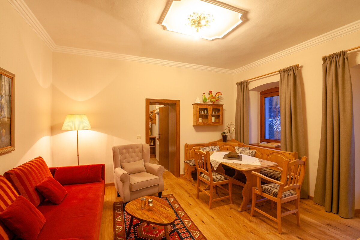 A comfortable living area features a red sofa and a beige armchair, complemented by a wooden dining table with chairs. Warm wood flooring adds to the inviting atmosphere, while a large window with curtains allows natural light to illuminate the space. A decorative chandelier hangs above.