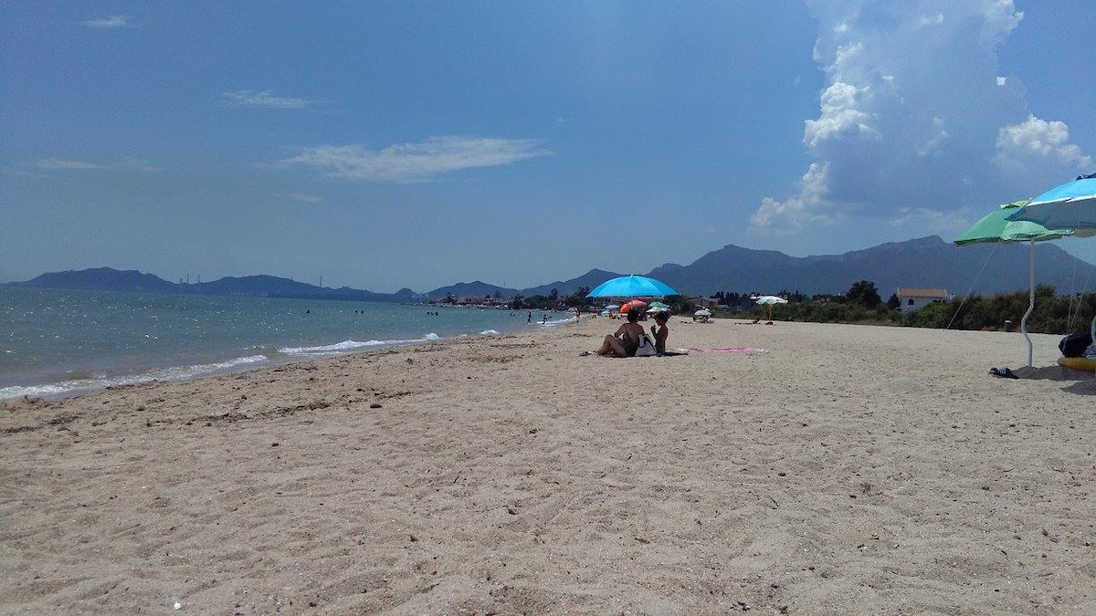 A sandy beach stretches along the shoreline, meeting the calm blue water. Sunbathers relax under colorful umbrellas. In the background, gentle mountains provide a natural backdrop, while a few people enjoy the sea. The sky is partly cloudy, enhancing the tranquil seaside setting.