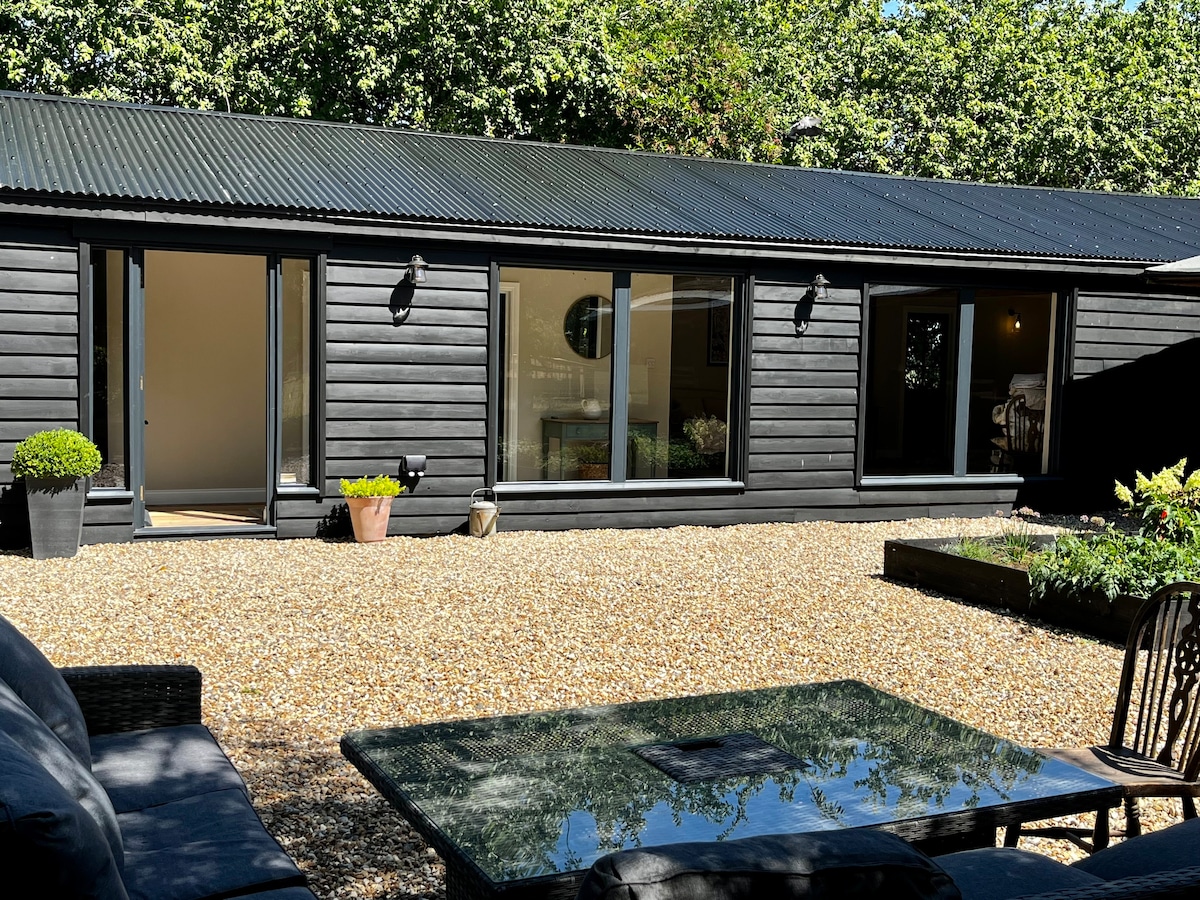 The exterior of the barn conversion features a sleek black wooden facade with large glass doors allowing natural light into the interior. A gravel patio area is equipped with outdoor seating and a glass table, surrounded by greenery and potted plants.
