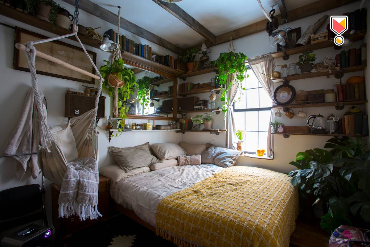 A guest room features a spacious bed adorned with multiple pillows and a light blanket. Shelves lined with books and plants create a cozy atmosphere. A hammock swing hangs nearby, and a window allows natural light to illuminate the room.
