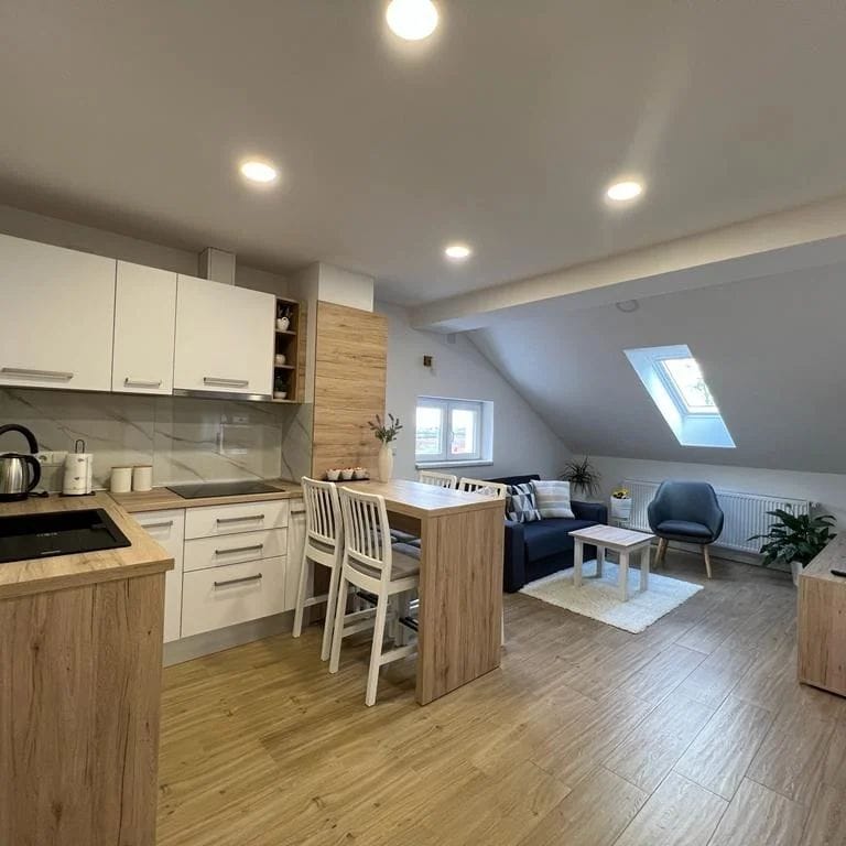 A modern kitchen and living area are captured in an open space, featuring light wood cabinetry and a central island with seating. Skylights allow natural light to illuminate the room, which includes a cozy seating area with a dark sofa and a light rug, along with potted plants.