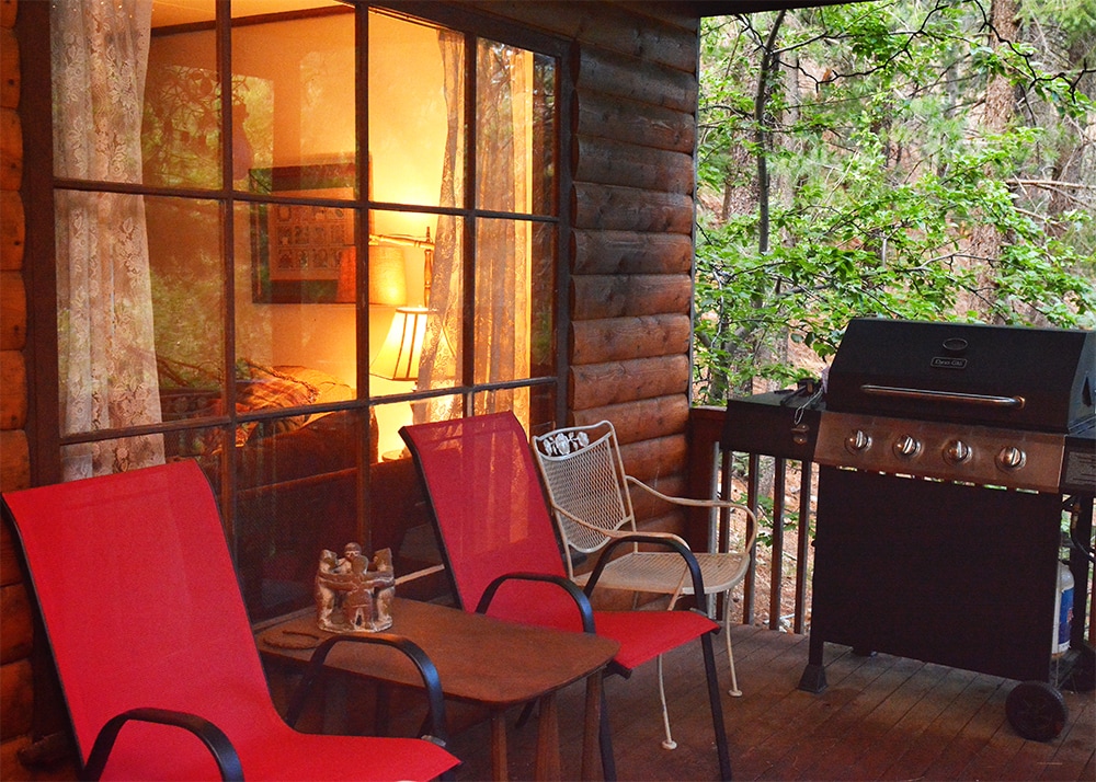 A porch is shown with two red chairs and a small table, creating a cozy seating area. A propane grill stands nearby, and a large window reveals a softly lit interior with a lamp. Surrounding trees provide a natural backdrop.