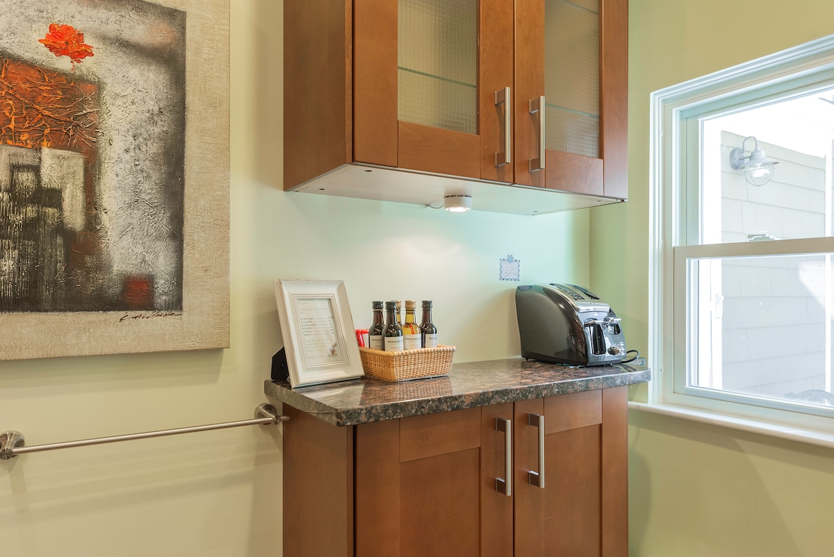 A kitchenette area is displayed with walnut-colored cabinetry and a granite countertop. A light basket holds various beverages, while a toaster is positioned beside a bright window, allowing natural light to fill the space. A modern piece of wall art adds a splash of color.