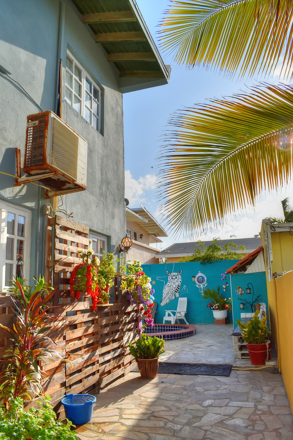 A colorful pathway leads to a private outdoor space, featuring a small seating area surrounded by tropical plants. The vibrant blue wall displays decorative art, while a few potted plants add to the inviting atmosphere. Sunlight illuminates the stone-paved ground.