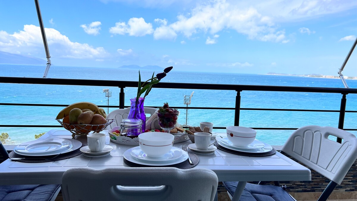 A dining table is set on a spacious balcony overlooking the sea, featuring plates, bowls, and a fruit basket. The vibrant blue waters and distant mountains create a scenic backdrop, enhancing the inviting outdoor dining experience.