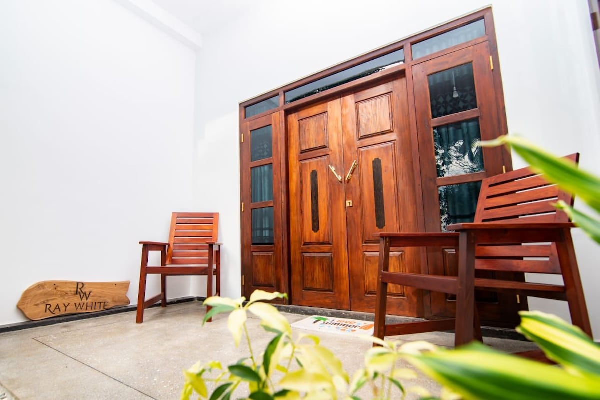 An inviting entrance features a pair of wooden chairs placed on either side of a sturdy door with elegant panels. A decorative sign is positioned along the floor, while lush green plants add a touch of nature to the calm ambiance.