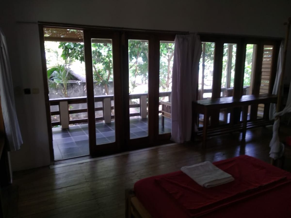 A spacious room features a sliding glass door opening onto a private terrace, surrounded by lush greenery. A wooden table is positioned near the door, and natural light filters through the curtains, illuminating the wooden flooring and warm-toned decor.