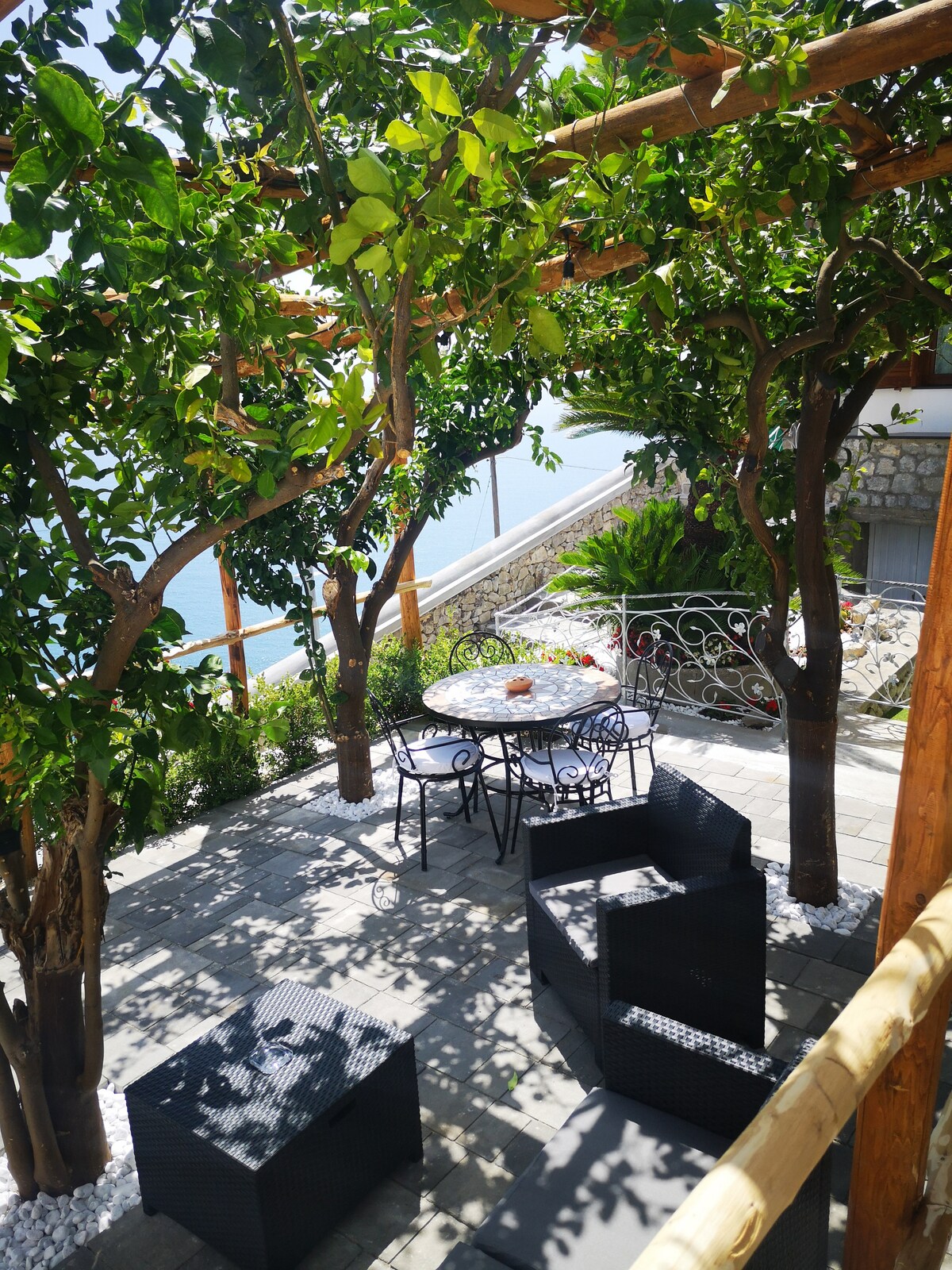 An outdoor seating area is surrounded by lush greenery, featuring a round table with four chairs beneath a lemon pergola. Two cozy armchairs are positioned nearby, providing scenic views of the sea and enhancing the relaxing ambiance of the space.
