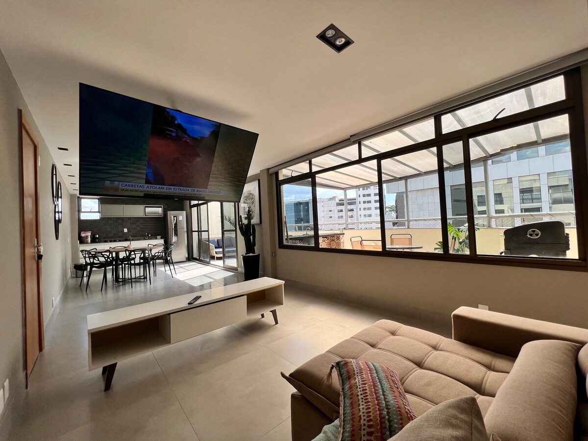A spacious living area is seen with large windows allowing for natural light. A smart TV mounted on the wall faces a comfortable sectional sofa. The open space leads to a terrace, which contains outdoor dining furniture visible through the windows.