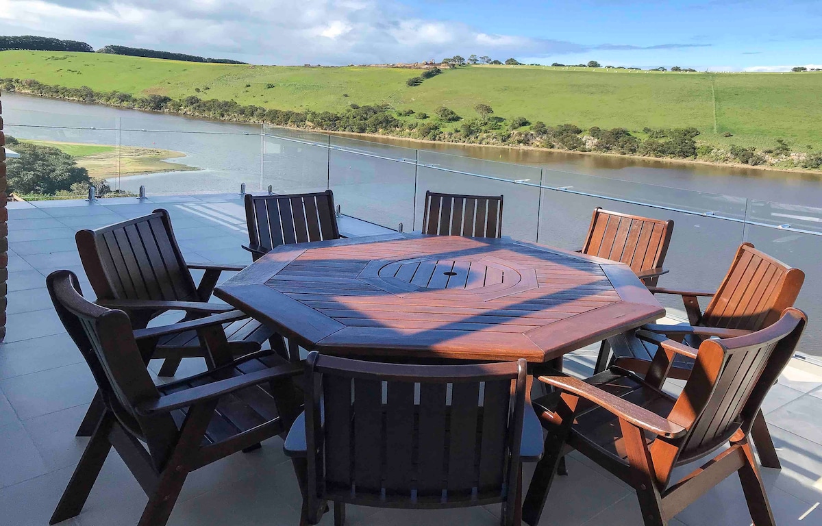 An octagonal wooden dining table is surrounded by six matching chairs on a spacious deck overlooking a serene river. A glass railing ensures an unobstructed view of the lush green landscape and calm waters beyond.