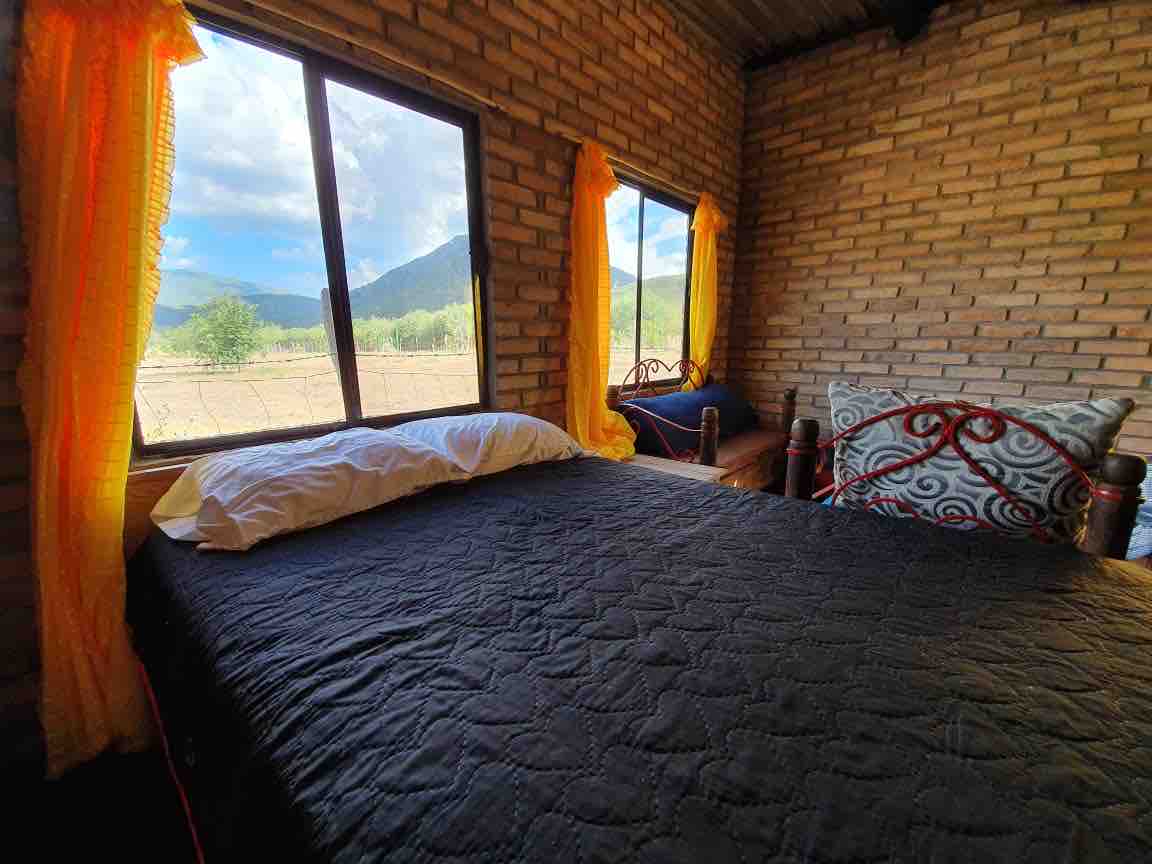 A cozy sleeping area features two beds with dark bedding, framed by large windows adorned with bright yellow curtains. Scenic views of the surrounding mountains and open fields are visible through the windows, allowing ample natural light to fill the space.