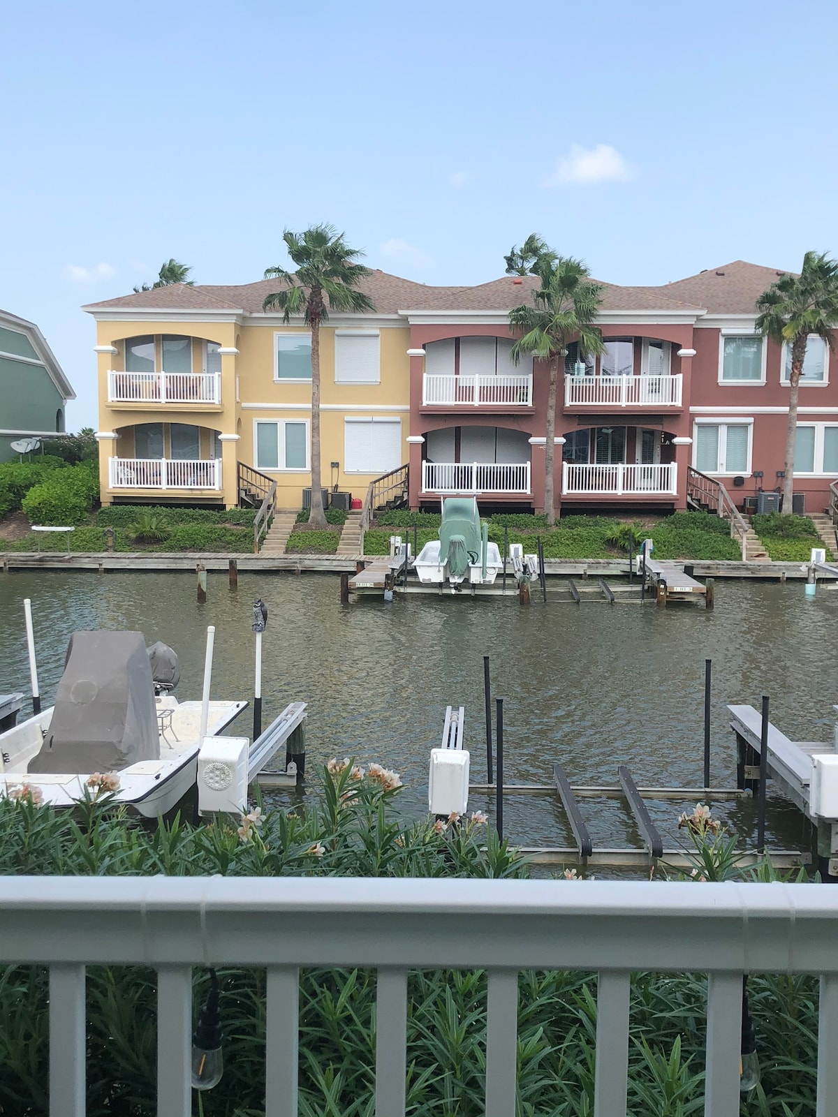 A view of a calm waterway is presented, with colorful villas lining the shore. The architecture reflects a blend of yellow and red hues. Boat docks are visible along the water's edge, surrounded by greenery and palm trees.