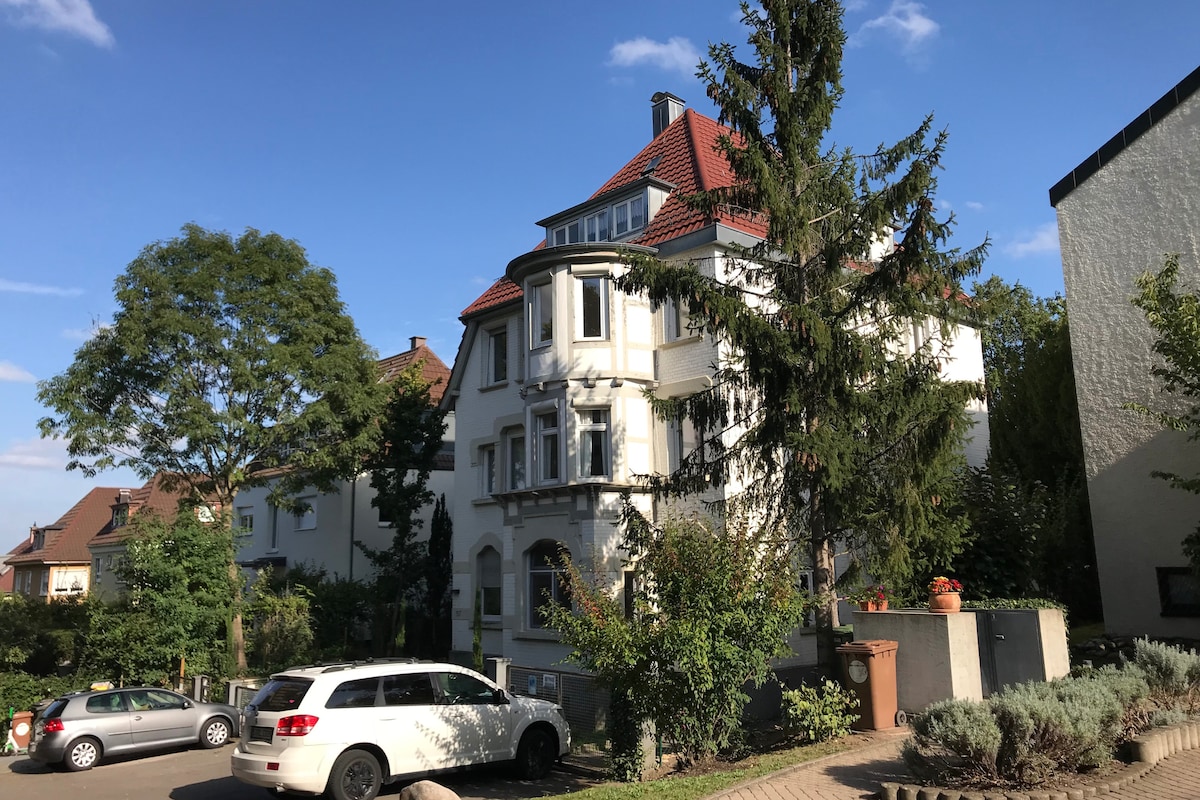 A three-story building with a distinctive architectural style is surrounded by greenery. A driveway leads to an entryway framed by trees and shrubs. Several cars are parked in the foreground, with the blue sky above displaying a few scattered clouds.