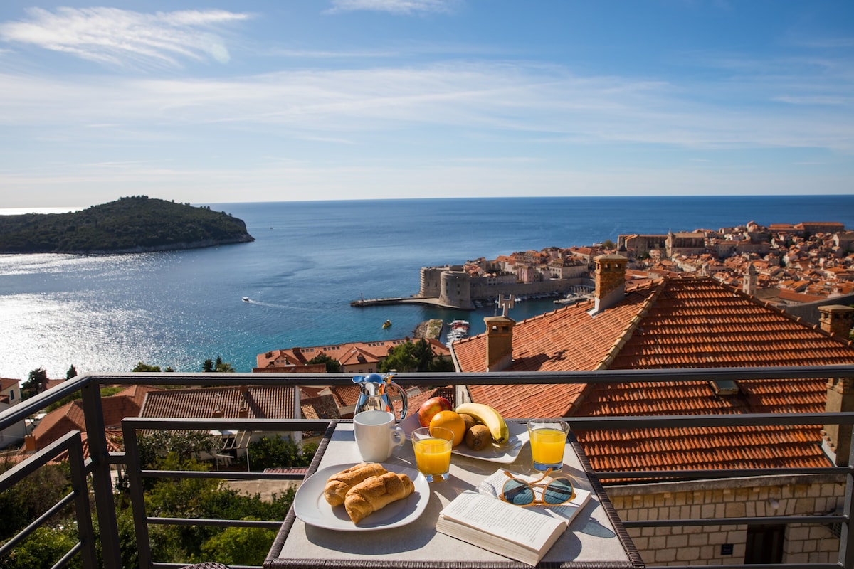 A balcony table is set with a light breakfast, featuring fresh fruit, a pastry, and beverages. Below, the coastline is visible, with historic buildings dotting the landscape and the water glistening in the sunlight.