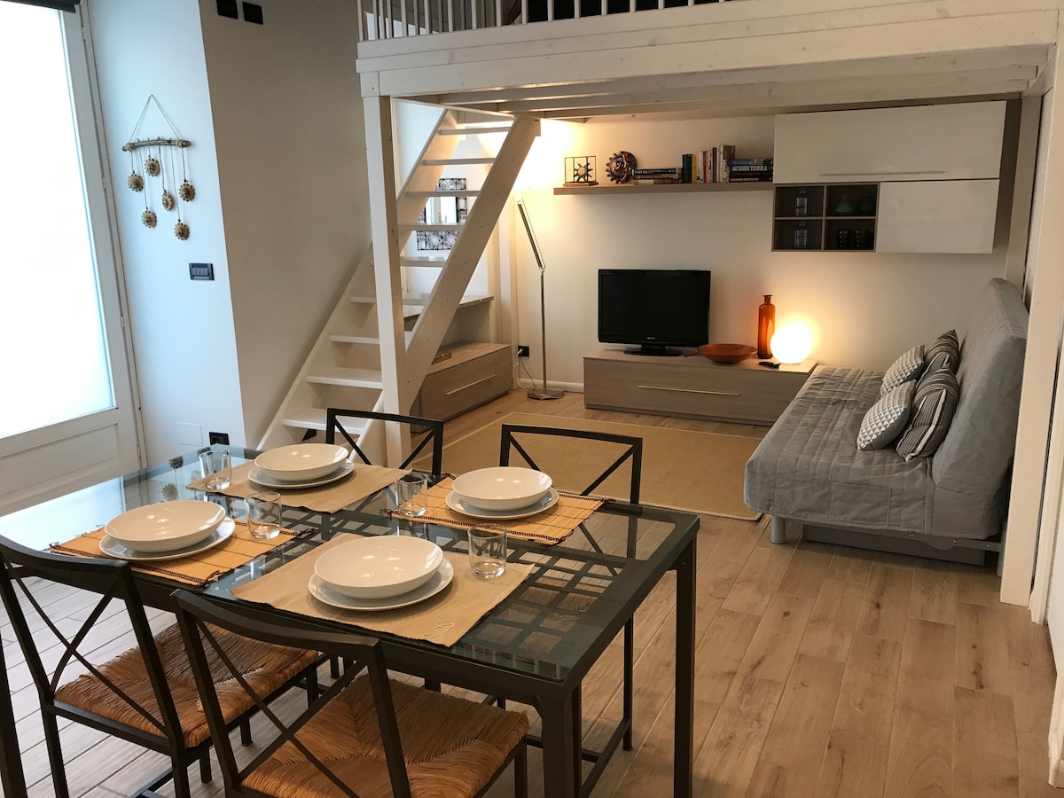 A spacious dining area features a glass table set for four, with plates and glasses arranged neatly. A comfortable sofa bed is positioned opposite a wall-mounted flat-screen TV, with a bookshelf adjacent to it. Natural light enters through large windows.