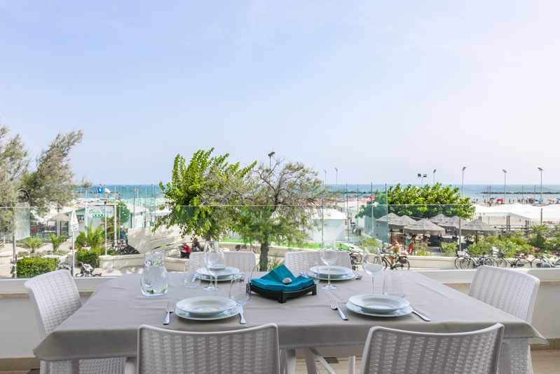 A balcony table is set for dining with white plates, glassware, and a turquoise menu holder. In the background, a beachfront scene is visible, showcasing a sandy beach, trees, and distant beach umbrellas under a clear sky.