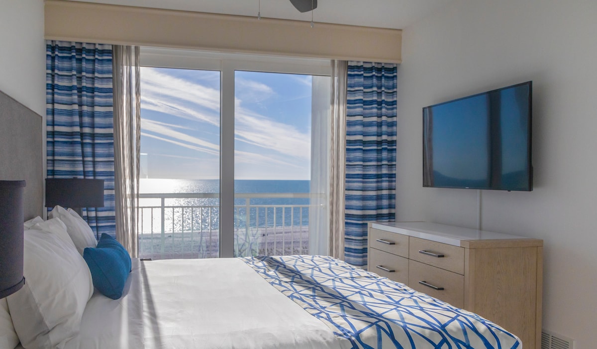 A bright bedroom features a spacious bed adorned with a blue and white patterned blanket. Large sliding doors provide a view of the ocean and sky, while sheer curtains frame the room. A modern dresser and wall-mounted TV are also included.