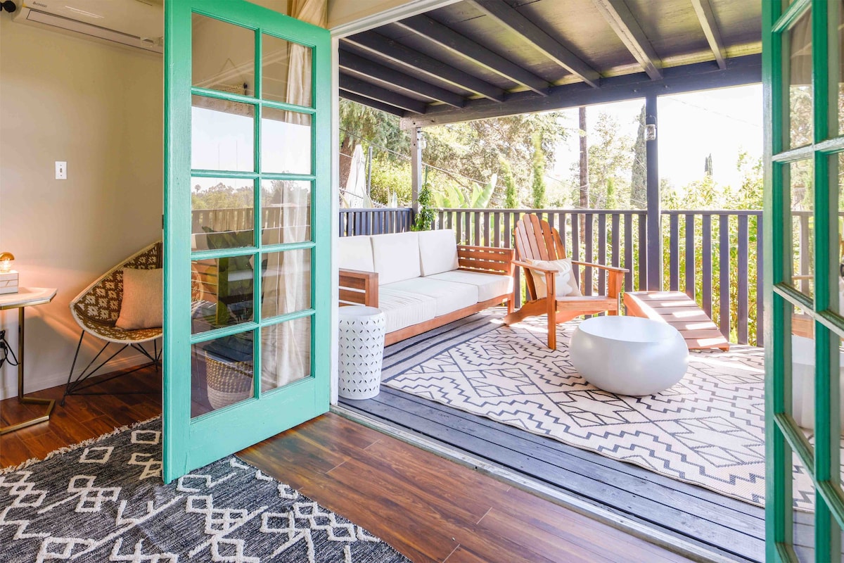 A serene outdoor seating area is depicted, featuring a cozy arrangement with a white sofa, two wooden chairs, and a circular table. A patterned rug lies beneath them, while lush greenery can be seen just beyond the railing, offering a peaceful view.
