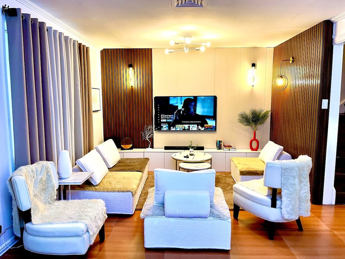 A comfortable living room is highlighted by soft seating arrangements, featuring several chairs and a couch with light-colored upholstery. A sleek wall-mounted television is positioned centrally, and soft lighting from modern fixtures adds a warm glow. Neutral tones and textures create an inviting space.