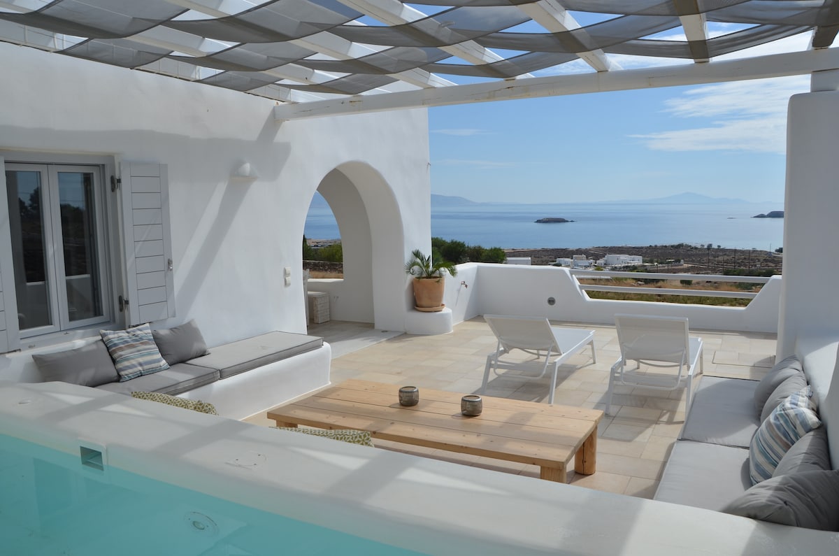 An open terrace features a wooden dining table with two decorative candles. Comfortable lounge furniture is positioned beside a serene swimming pool. The view extends towards the sea, with gentle waves visible under a clear sky, enhancing the tranquil outdoor space.