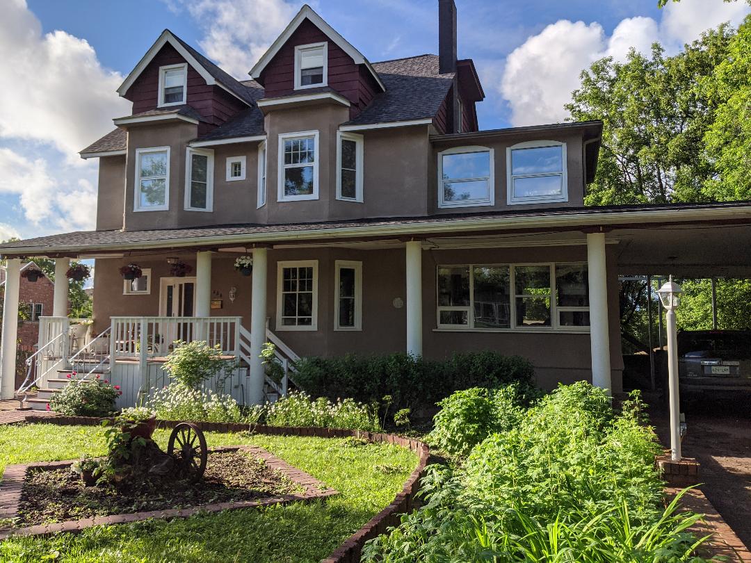 The exterior of the Little Mansion features a charming three-story structure with a gabled roof and multiple windows. A welcoming porch with steps leads to the entrance, surrounded by lush greenery and blooming plants, enhancing the home’s historic character.