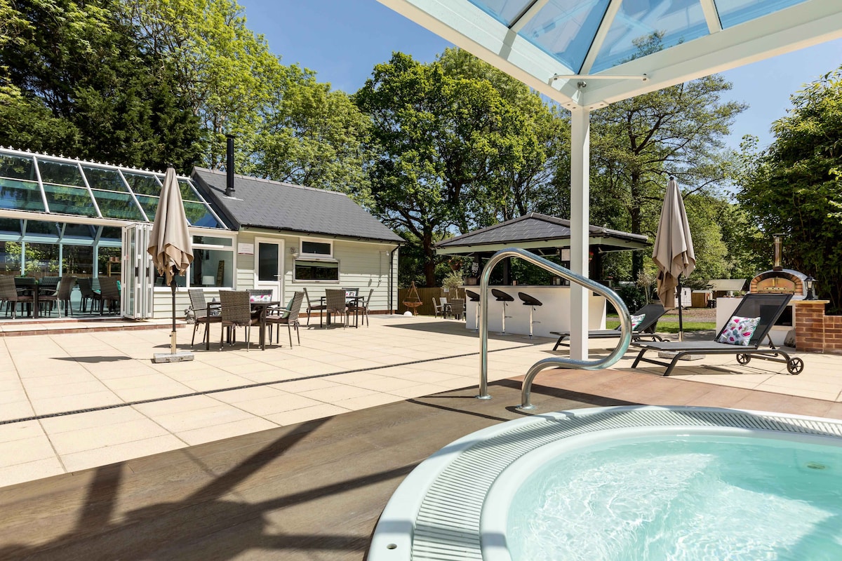 The image captures a sunny terrace area featuring a hot tub in the foreground. Beyond, modern outdoor seating arrangements are visible, along with shaded tables and umbrellas. The well-kept grounds are surrounded by greenery, creating a serene and relaxing environment for guests.