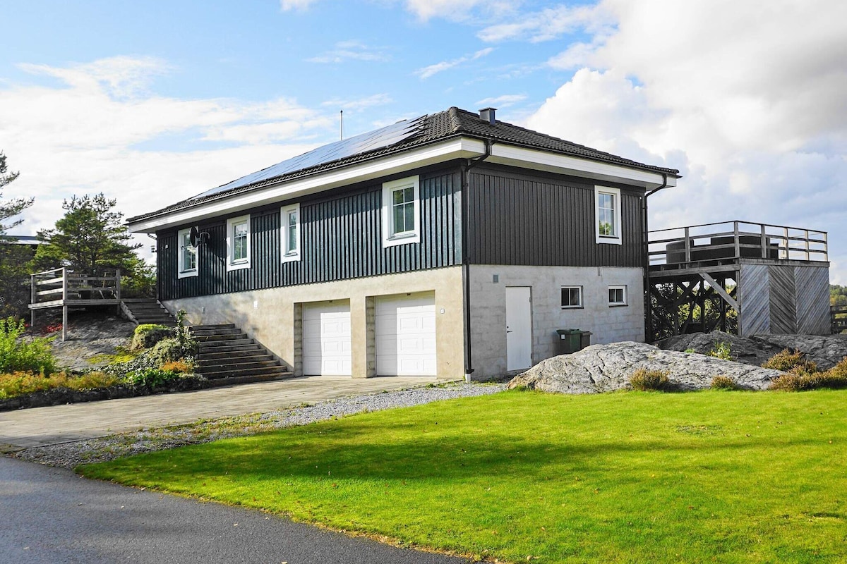 A modern two-story holiday home is set on a hilly nature plot, featuring a green and gray exterior. A white garage is visible beneath the house, along with a staircase leading to the upper level, which has a deck offering views of the surrounding landscape.
