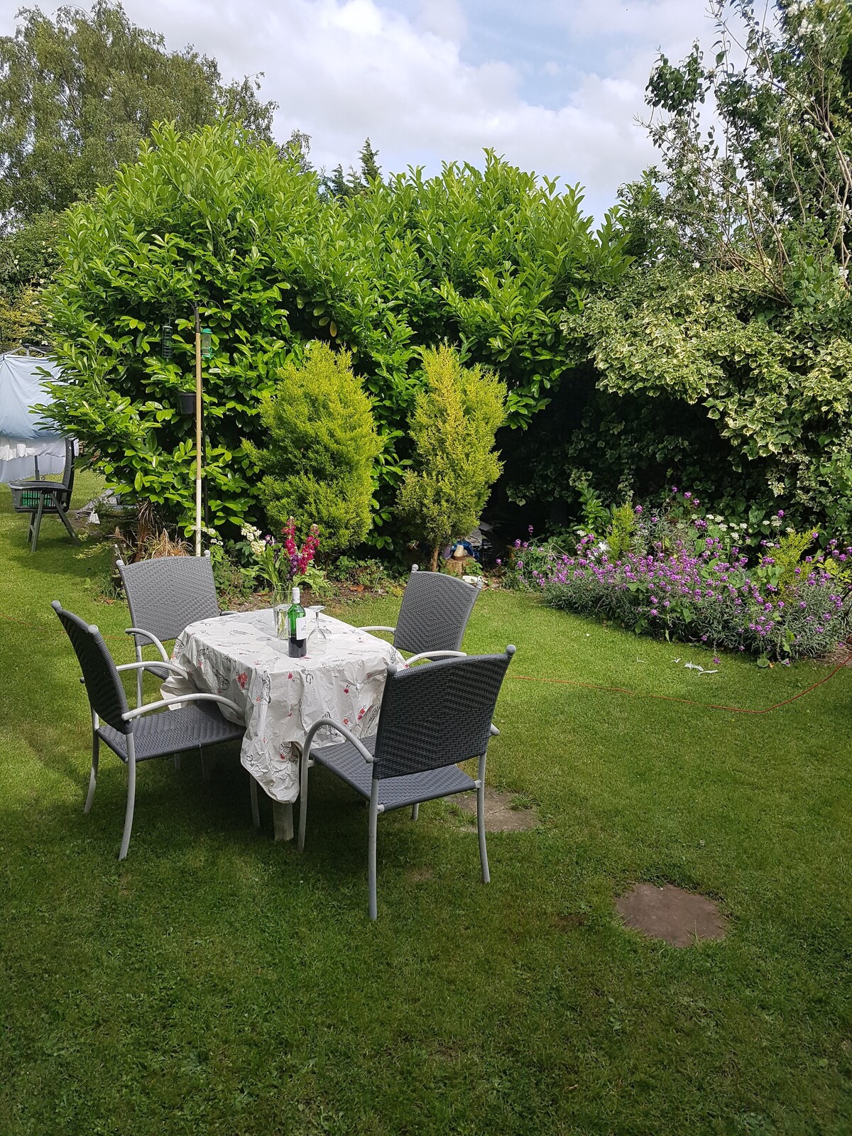 A garden area features a round table surrounded by four gray chairs, set on a manicured lawn. The table is adorned with a vase of fresh flowers. Lush greenery and blooming plants frame the space, providing a serene outdoor setting.