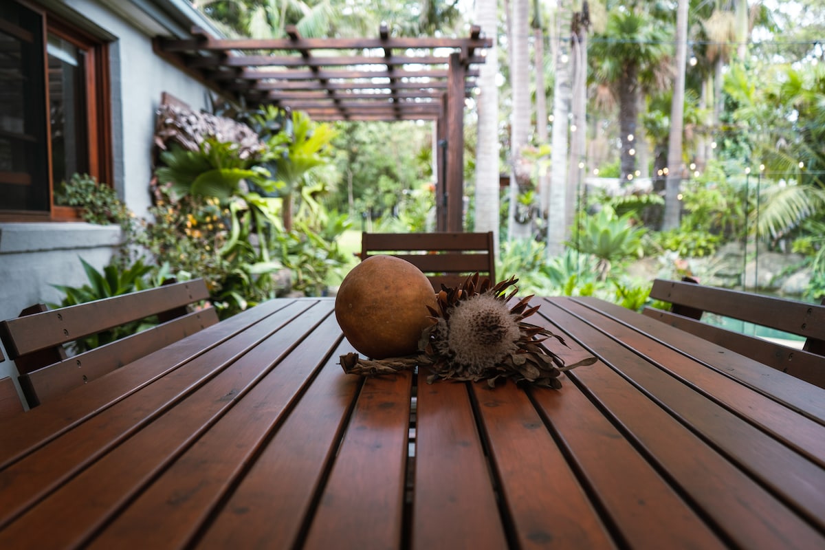 A wooden dining table, made of slatted timber, is set outdoors. Centered on the table are tropical fruits and foliage, with a background of lush greenery and landscaped garden elements visible. The setting conveys a tranquil outdoor dining space surrounded by nature.