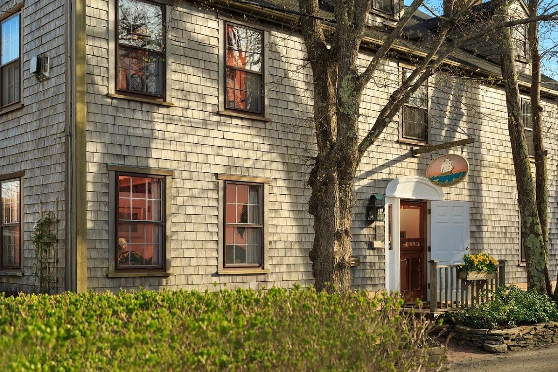 The exterior of the historic bed and breakfast showcases traditional shingle siding and large windows framed by greenery. A welcoming entrance is centered beneath a decorative sign, surrounded by flowering plants and a stone pathway leading to the door.