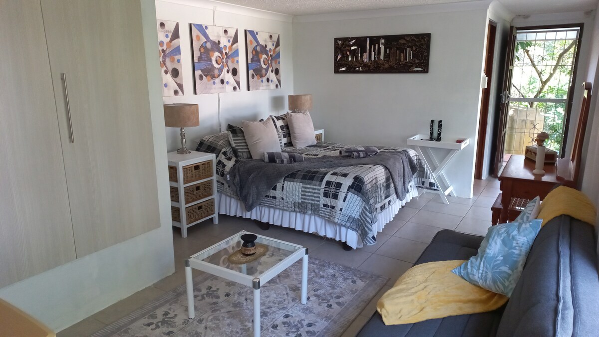 A cozy bedroom space features a spacious bed adorned with textured pillows and a quilt. Two bedside lamps provide soft lighting. A small table sits in front of the bed, complemented by a comfortable sofa. Artwork adorns the walls, enhancing the room's inviting ambiance.