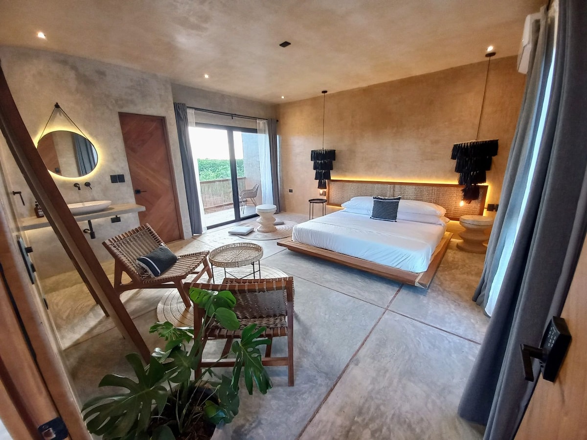 A spacious bedroom is featured with a large bed positioned against a textured wall. Natural light enters through sliding glass doors leading to a private balcony. Simple wood furniture, including a lounge chair and a small table, complements the serene atmosphere, alongside a small indoor plant.