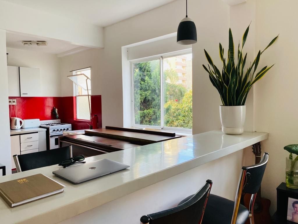 The kitchen area features modern cabinetry and vibrant red backsplash, complemented by a window overlooking greenery. A sleek countertop is adorned with a laptop and notebook, while a potted plant adds a touch of freshness. Black bar stools provide seating near the countertop.