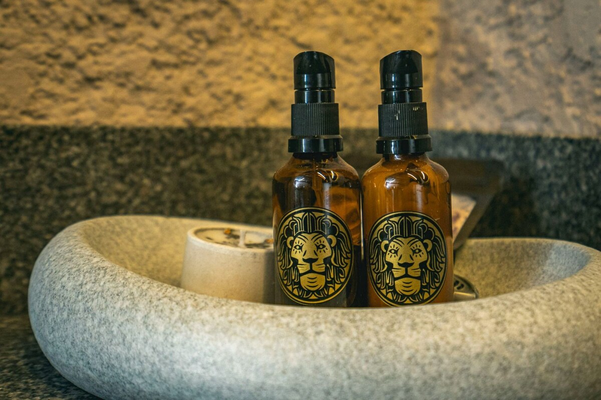 Two amber bottles with lion-themed labels are positioned on a textured stone dish, accompanied by a small, light-colored candle. The composition is set against a speckled granite surface, emphasizing a neat and organized presentation.