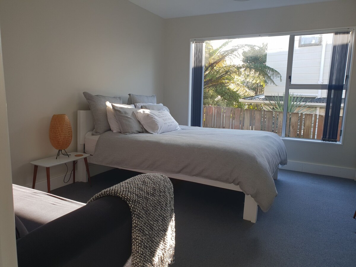 A spacious sleeping area features a comfortable queen bed covered with light grey linen. A cozy sofa with a textured throw is visible in the foreground. Large windows allow natural light and views of greenery from outside, enhancing the bright atmosphere.