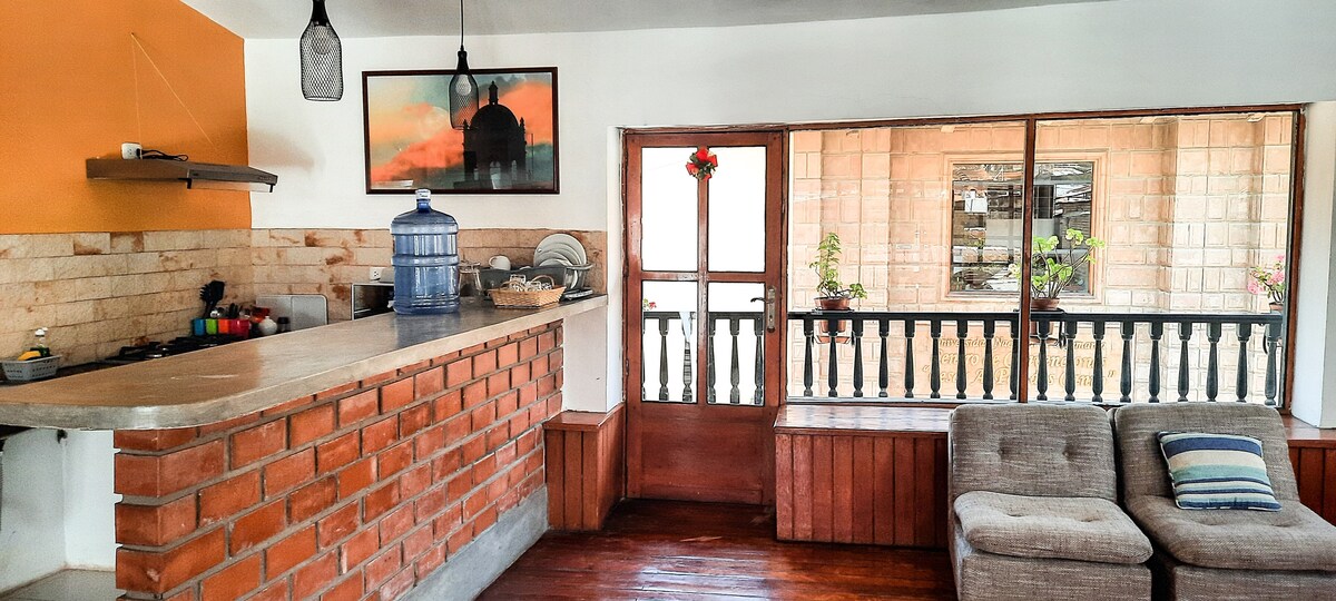 A bright, open kitchen area features a unique brick bar with seating and a modern cooking setup. Large windows reveal a view of an outdoor balcony adorned with potted plants. Natural light fills the space, creating a warm, inviting environment.
