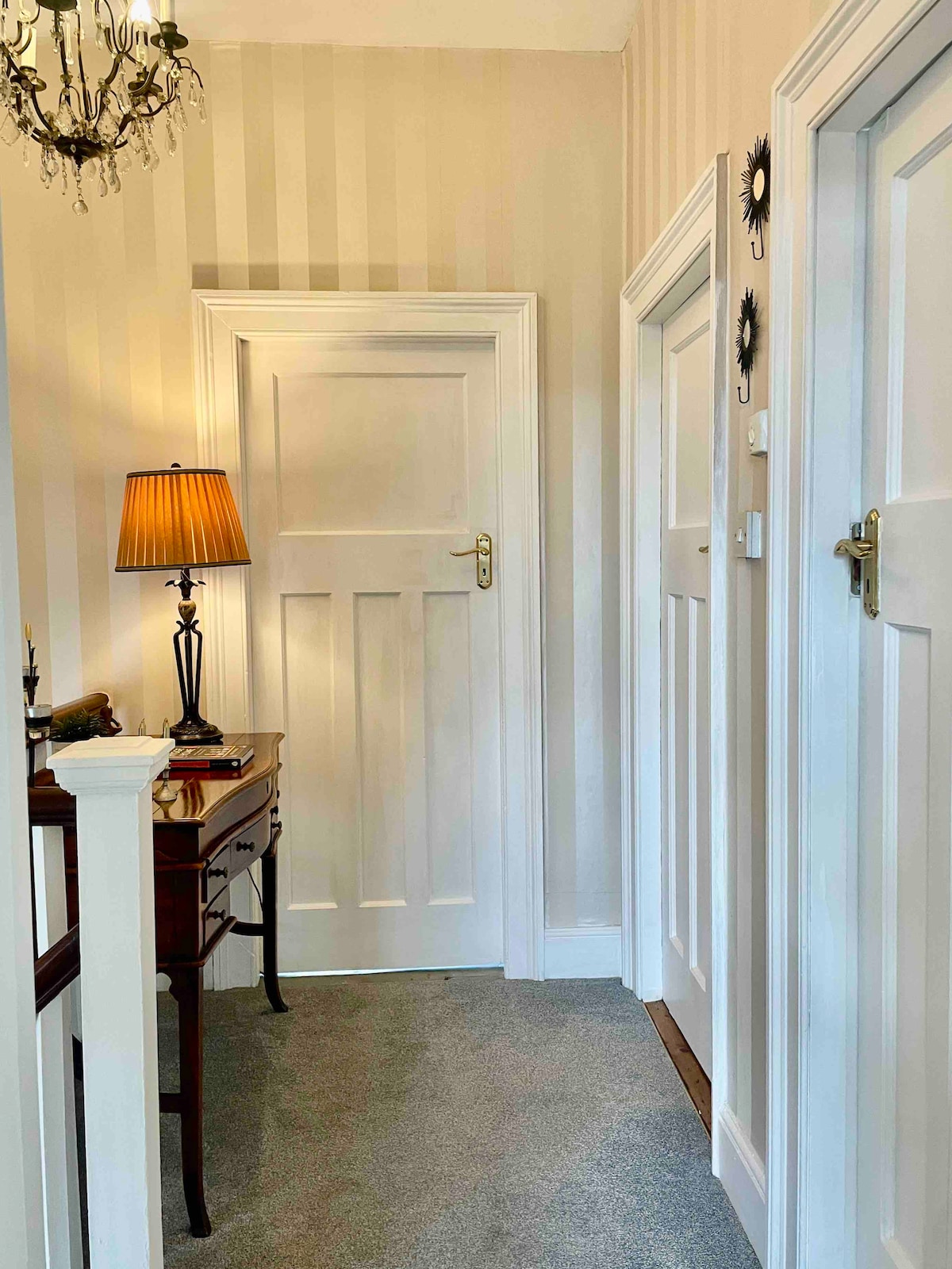 A hallway featuring soft striped wallpaper and plush gray carpet. A vintage wooden table is positioned on one side, accompanied by a decorative lamp providing soft illumination. Two closed doors are visible at the end of the corridor, contributing to a serene and organized atmosphere.