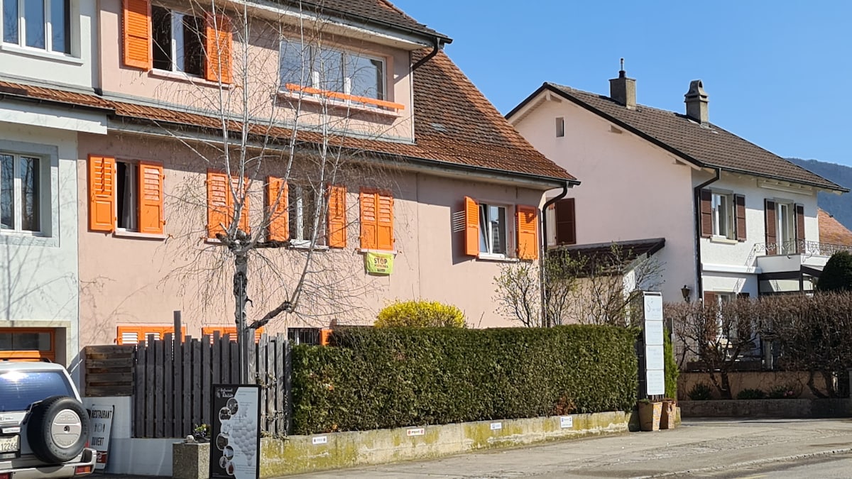 A well-maintained building with a light pink exterior is featured, accented by bright orange window shutters. A manicured hedge runs along the front, with a small tree visible to the left. The setting includes nearby residential structures under a clear blue sky.