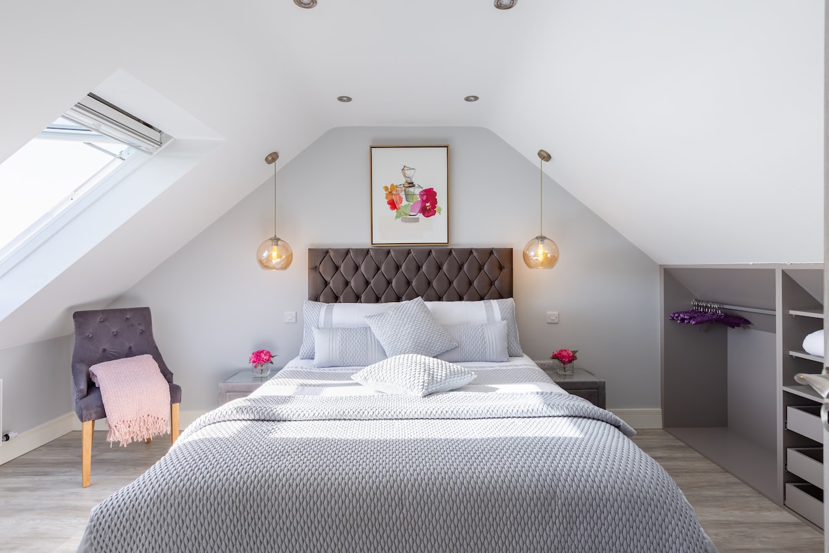 A serene bedroom with slanted ceilings features a spacious bed adorned with a neutral blanket and decorative pillows. Beside the bed, stylish pendant lights offer illumination. A chair is positioned in the corner, complemented by a pink throw. Built-in storage provides organizational convenience.
